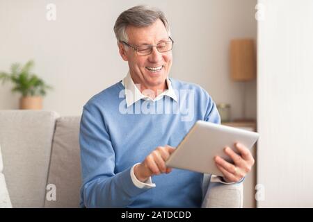 Senior Man With Tablet Computer Browsing Internet Sitting On Sofa Stock Photo