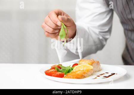 Male Chef Plating Fish Dish Standing In Kitchen, Cropped Stock Photo