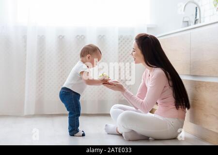 Cute boy giving apple to mom sitting on kitchen floor Stock Photo