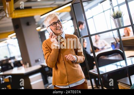 Senior businesswoman using mobile phone at office while other people having meeting in background Stock Photo