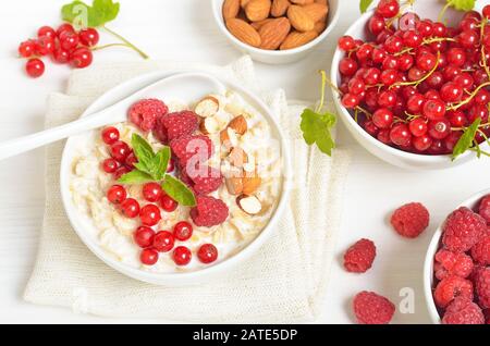 Oatmeal porridge with raspberries, red currant and nuts in bowl on white table. Fresh berries in bowl. Top view, flat lay Stock Photo