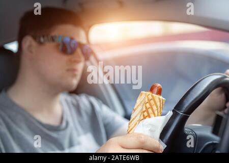 Adult male caucasian person holding hot dog fast food snack while driving car on sunny day. Men eating food while driving steering wheel. Distracted Stock Photo