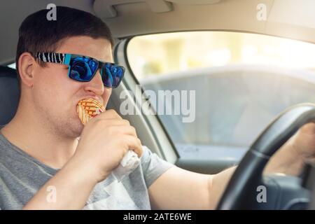 Adult male caucasian person eating hot dog fast food snack while driving car on sunny day. Men hsving food while driving steering wheel. Distracted Stock Photo