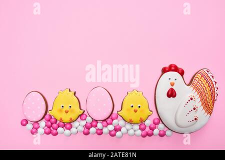 From above view of glazed cookies in shape of lovely chicken, two yellow chicks and eggs, isolated on pink background. Close up of cute pastry and small chocolate balls in sugar shell in row. Stock Photo