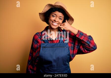 Young African American afro farmer woman with curly hair wearing apron and hat smiling doing phone gesture with hand and fingers like talking on the t Stock Photo