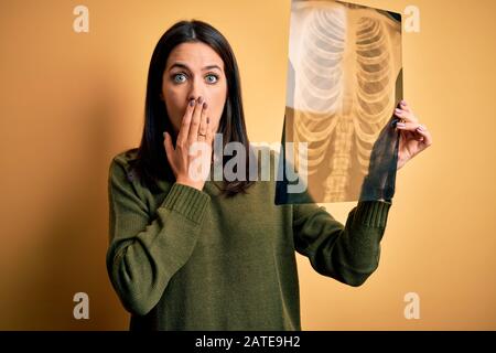 Young brunette woman with blue eyes holding xray of lungs over yellow background cover mouth with hand shocked with shame for mistake, expression of f Stock Photo