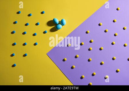 From above view of chocolate peanut balls in colorful crispy sugar shell and three blue eggs isolated on yellow and purple geometric background. Lovely delicious candies lying on table, shadows. Stock Photo