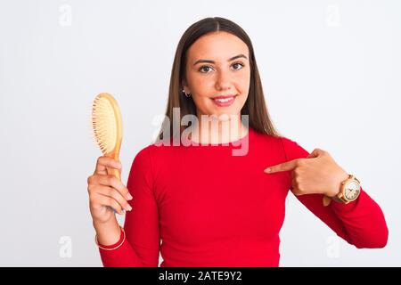 Young beautiful girl holding hair comb standing over isolated white background with surprise face pointing finger to himself Stock Photo