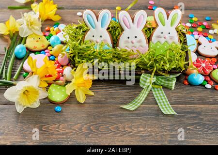 Easter ginger glazed cookies in shape of animals, eggs, three bunnies in box with fake grass, daffodils isolated on wooden background. Peanut chocolate balls in sugar shell. Concept of holidays. Stock Photo