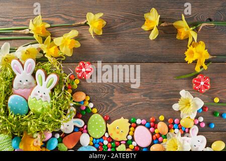 Peanut chocolate balls in sugar shell and daffodils isolated on wooden background. Easter ginger glazed cookies in shape of animals, colorful eggs and bunnies in green nest. Concept of holidays. Stock Photo