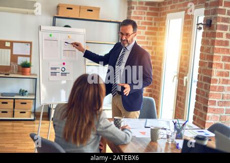 Two middle age business workers smiling happy and confidentt working together with smile on face. Man standing doing work presentation using board and Stock Photo
