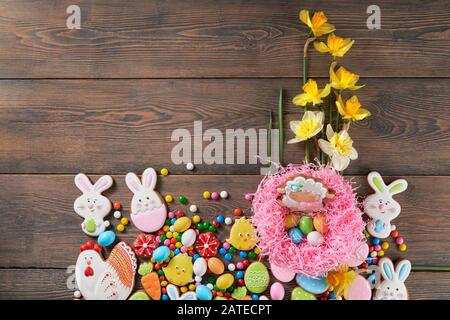 From above view of peanut chocolate balls in crispy sugar shell and daffodils isolated on wooden background. Easter ginger glazed cookies in shape of animals, colorful eggs and sheep in pink nest. Stock Photo