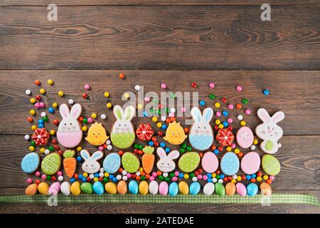 Big and small colorful peanut chocolate balls in crispy sugar shell and tartan ribbon isolated on wooden background. Easter ginger glazed cookies in shape of bunnies, flowers, carrots and eggs in row. Stock Photo