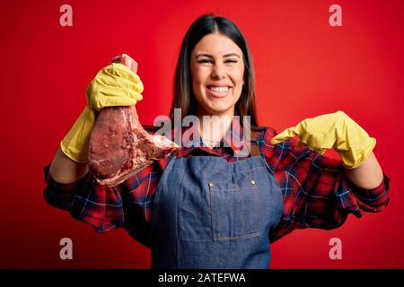 Young butcher woman holding fresh raw beef meat stake over red background with surprise face pointing finger to himself Stock Photo