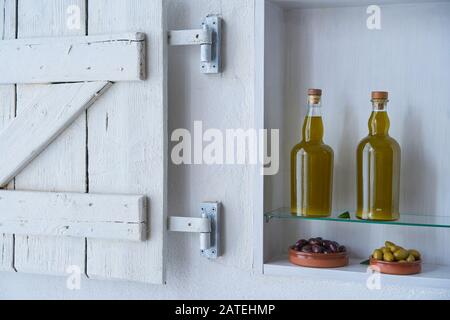 Pure and stylish shelf with a white wooden shabby shutter in a country house with olives in bowls and olive oil in glass bottles, pure and white Stock Photo