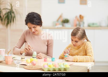 Candid portrait of mother and daughter painting Easter eggs pastel colors sitting at table in cozy kitchen interior, copy space Stock Photo