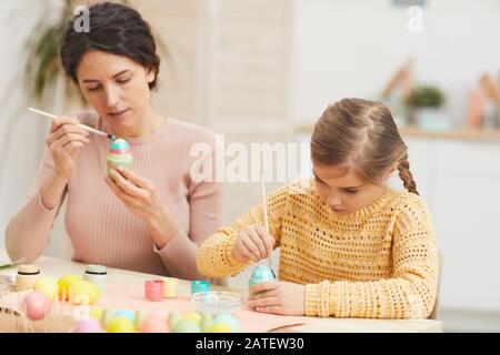 Portrait of mother and daughter painting Easter eggs together sitting at table in cozy kitchen interior, copy space Stock Photo