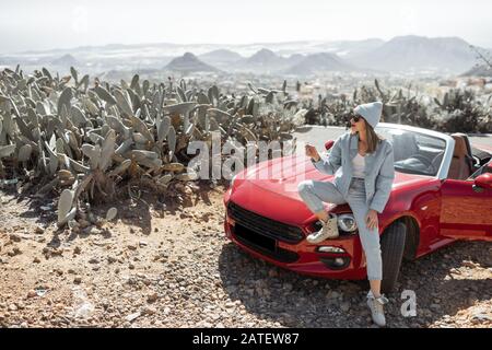 Young woman dressed casually with hat enjoying road trip on the island, standing with phone near the convertible car on the roadside Stock Photo
