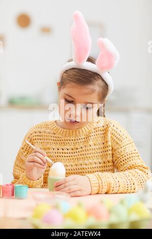 Vertical portrait of cute little girl painting Easter eggs while wearing bunny ears in cozy kitchen interior, copy space Stock Photo