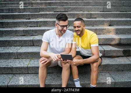 Portrait of happy gay couple spending time together and using a digital tablet while sitting on stairs. Lgbt and love concept. Stock Photo