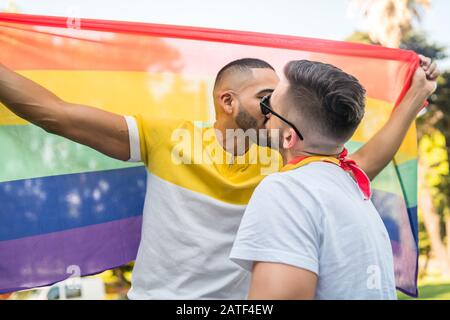 Portrait of young gay couple kissing and showing their love with rainbow flag in the stret. LGBT and love concept. Stock Photo