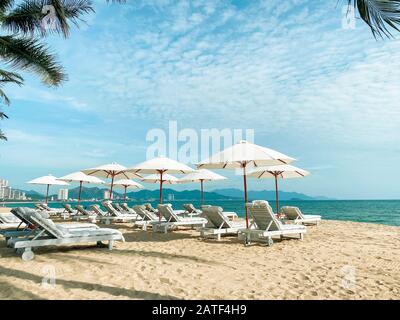 White soft sun loungers on the shore of a sandy sea beach. Tropical resort, morning sun, white clouds in the blue sky, view of the sea, mountains and Stock Photo