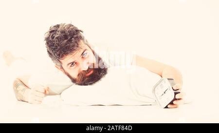 Man unhappy in morning, wants to turn off alarm clock, white background. Hipster with beard and mustache awake because of alarm clock. Morning wake up concept.Guy with tired face holds alarm clock. Stock Photo