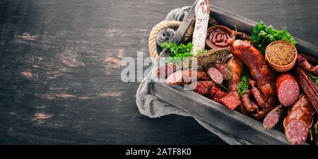 Assortment of sausages and snacks in a wooden box. Sausage Fuet, salami, paperoni. On a black background. Top view. Free space for your text. Stock Photo
