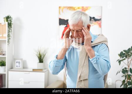 Tired senior man in blue shirt touching his temples while feeling headache Stock Photo