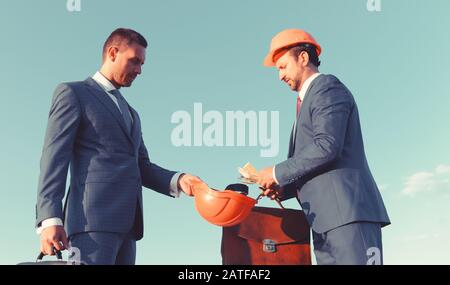 Engineers and cash on blue sky background. Bribery and business bargain concept. Architects with serious faces in suits and helmets share budget. Builder with case puts money into colleagues hardhat Stock Photo