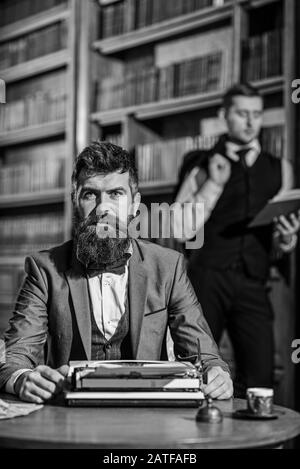 Bearded writer with concentrated face typing on a typewriter in front of library, near his friend, poet or researcher. Writer works with typewriter, defocused Stock Photo