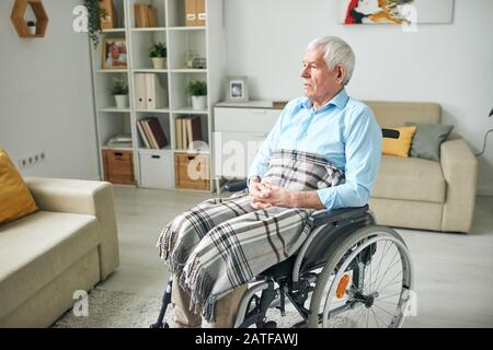 Sad and serene senior disable man sitting in wheelchair by couch at home Stock Photo