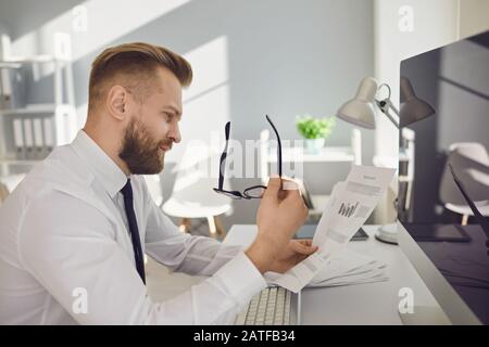 Serious busy businessman reads working documents sitting at a table with a computer in the office. Stock Photo