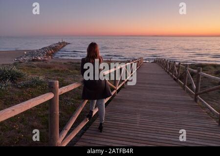 Woman looking sunset in Weathered lumber path leading to calm sea during beautiful sunset in countryside in Cabopino, Artola dunes. Marbella Stock Photo