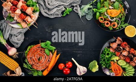 Assortment of baked barbecue meat. Sausages, skewers, fresh vegetables. Tomatoes, onions, garlic. On a wooden background. Copy space. Stock Photo