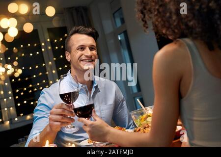 Romantic Date. Young multiethnic couple at home sitting at table eating dinner cheers with glasses of wine talking smiling cheerful Stock Photo