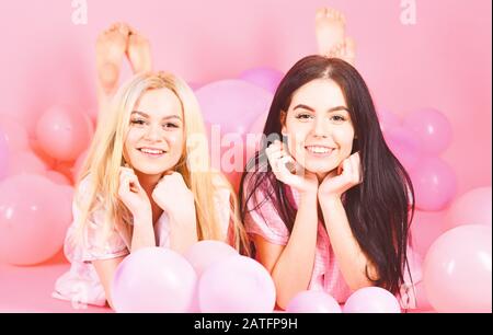 Girls lay on belly near balloons, pink background. Gossip concept. Blonde and brunette on smiling faces have fun at domestic bedroom party. Sisters or best friends in pajamas at girlish pajamas party. Stock Photo