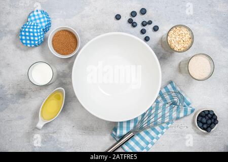 Baking background, preparation of sweet pastry. Empty white bowl, milk, sugar, flour, oil blueberries and paper muffin cups Stock Photo