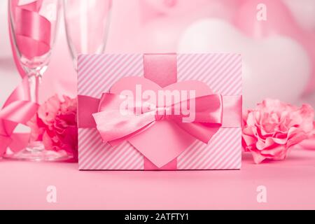 Valentine Day gift in a box wrapped in striped paper and tied with silk ribbon bow and heart shaped greeting card on pink balloons background with cop Stock Photo