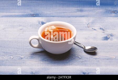 Process of black tea brewing in mug. Mug filled with boiling water, teabag and spoon on blue wooden texture background. Tea brew concept. Mug filled with hot water and dipped bag of black tea. Stock Photo