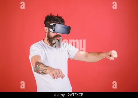 Virtual racing concept. Hipster on confident face driving bike on high speed in virtual reality. Man with beard in VR glasses driving motorbike, red background. Guy play racing game in VR. Stock Photo