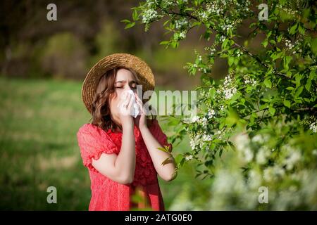 Young girl blowing nose and sneezing in tissue in front of blooming tree. Seasonal allergens affecting people. Beautiful lady has rhinitis. Stock Photo