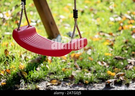 Empty red swing on children playground in the park in autumn season. Missing child. Lonely swing in sunny autumn day. Stock Photo