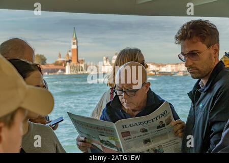A man reading a newspaper standing next to other passengers on a Vaporetto or water bus service on The Venice Lagoon, on route to Venice ,Italy