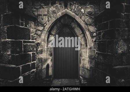 Scary pointy wooden door in an old and wet stone wall building with cross, skull and bones at both sides in black and white. Concept mystery, death Stock Photo