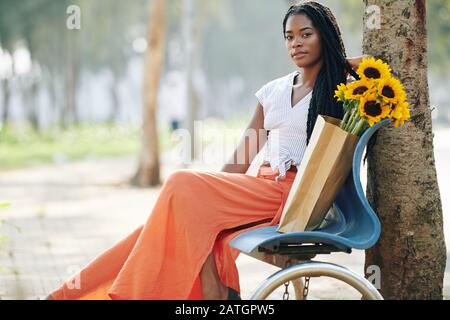 Beautiful young Black woman with braided hair sitting on bench in park with bouquet of sunflowers Stock Photo