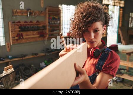 Young concentrated female carpenter with curly hair holding wooden plank and estimating its length before sawing Stock Photo