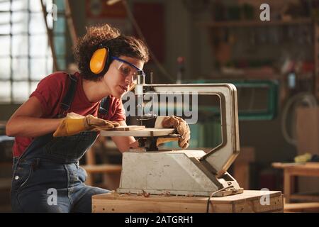 Young concentrated woman in protective eyewear and earmuffs making holes in wooden plank with drill press Stock Photo