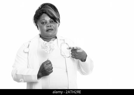 Studio shot of fat black African woman doctor holding and pointing at heart shaped lollipop