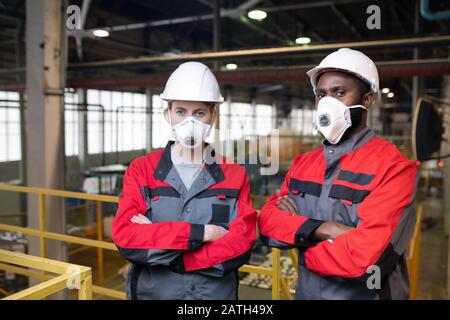 Portrait of serious workers in respiratory masks and hardhats standing with crossed arms on frame bridge in factory shop Stock Photo